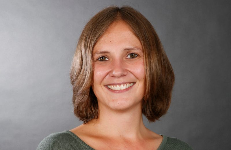 Businessfoto einer jungen Frau mir braunen mittellangen Haaren, freundlichem offenen Lächeln. Fotostudioaufnahme mit grauem Hintergrund in Göttingen