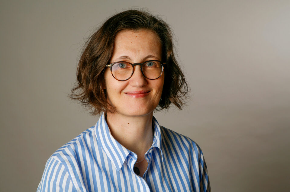 Professionelles Porträt einer Frau mit Brille, gestreiftem Hemd und natürlichem Ausdruck vor hellem Studiohintergrund.