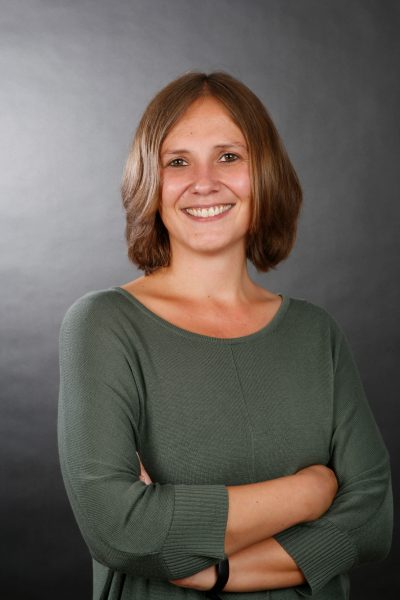 Businessfoto einer jungen Frau mir braunen mittellangen Haaren, freundlichem offenen Lächeln. Fotostudioaufnahme mit grauem Hintergrund in Göttingen