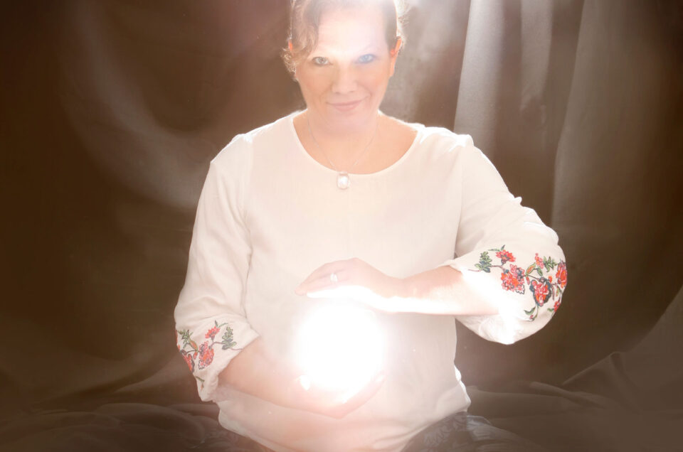 Auf diesem Foto wird ein "Seelenfoto" einer Frau gezeigt. Sie hält einen Lichtball in der Hand und einer leuchtet hinter ihr. Es ist eine Studioaufnahme mit schwarzem Hintergrund der weiche Falten wirft. Es ist ein mystisches Bild, was Ruhe und Kraft ausstrahlt.