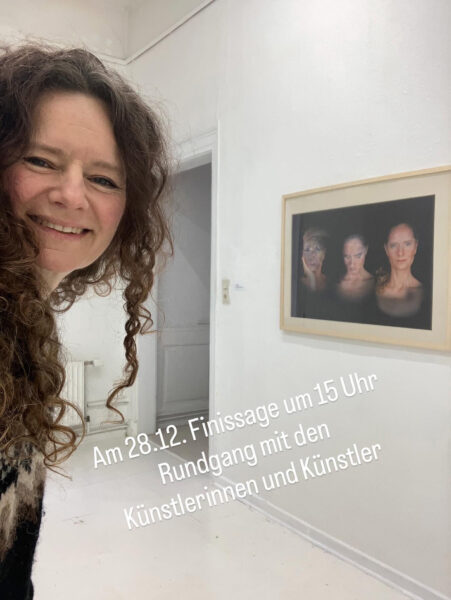 Frau lächelt in einem weißen Ausstellungsraum neben einer gerahmten Fotografie an der Wand; im Bild steht der Text „Am 28.12. Finissage um 15 Uhr Rundgang mit den Künstlerinnen und Künstlern“. Kunstausstellung vom BBK.