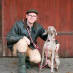 Ein Mann mit Brille und Schiebermütze kniet neben einem eleganten grauen Hund, vermutlich ein Weimaraner, vor einer rustikalen roten Holzwand. Der Hund trägt ein Halsband mit Leine und blickt aufmerksam zur Seite, während sein Besitzer entspannt lächelt. Die natürliche Umgebung und das weiche Licht schaffen eine authentische und ruhige Atmosphäre. Professionelle Tier- und Mensch-Hund-Fotografie von Sonja Mehner in Göttingen.